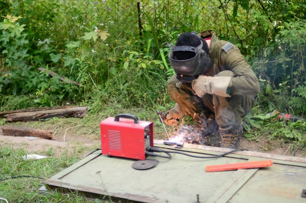 Kahverengi üniformalı yaşlı kaynakçı, kaynak maskesi ve kaynakçı derisi, kaynak makinesi olan metal kapılar açık havada, mavi kıvılcımlar yanlarda uçuşuyor.