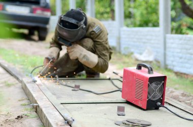Kahverengi üniformalı yaşlı kaynakçı, kaynak maskesi ve kaynakçı derisi, kaynak makinesi olan metal kapılar açık havada, mavi kıvılcımlar yanlarda uçuşuyor.