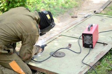 Kahverengi üniformalı yaşlı kaynakçı, kaynak maskesi ve kaynakçı derisi, kaynak makinesi olan metal kapılar açık havada, mavi kıvılcımlar yanlarda uçuşuyor.