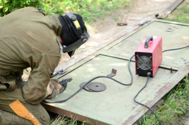 Kahverengi üniformalı yaşlı kaynakçı, kaynak maskesi ve kaynakçı derisi, kaynak makinesi olan metal kapılar açık havada, mavi kıvılcımlar yanlarda uçuşuyor.
