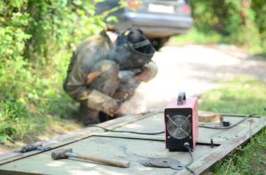 Kahverengi üniformalı yaşlı kaynakçı, kaynak maskesi ve kaynakçı derisi, kaynak makinesi olan metal kapılar açık havada, mavi kıvılcımlar yanlarda uçuşuyor.