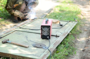 Kahverengi üniformalı yaşlı kaynakçı, kaynak maskesi ve kaynakçı derisi, kaynak makinesi olan metal kapılar açık havada, mavi kıvılcımlar yanlarda uçuşuyor.