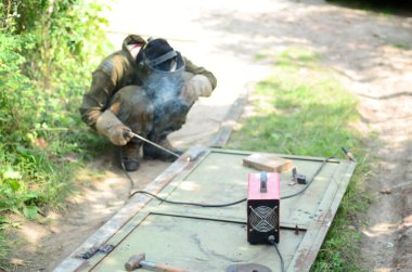Kahverengi üniformalı yaşlı kaynakçı, kaynak maskesi ve kaynakçı derisi, kaynak makinesi olan metal kapılar açık havada, mavi kıvılcımlar yanlarda uçuşuyor.
