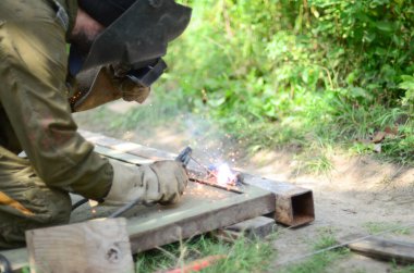 Kahverengi üniformalı yaşlı kaynakçı, kaynak maskesi ve kaynakçı derisi, kaynak makinesi olan metal kapılar açık havada, mavi kıvılcımlar yanlarda uçuşuyor.