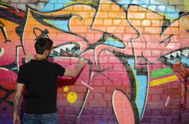 Sırt çantası ve gaz maskesi takan genç grafiti sanatçısı, tuğla duvara pembe tonlarda grafiti yapıyor. Sokak sanatı ve çağdaş boyama süreci. Gençlik alt kültüründe eğlence