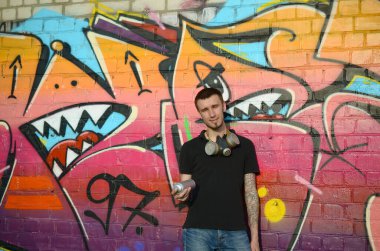 Beyaz beyaz grafiti sanatçısı siyah tişörtlü, sprey sprey boyalı tuğla duvarında renkli pembe grafitilerin yanında. Sokak sanatı ve çağdaş boyama süreci. Gençlik alt kültüründe eğlence