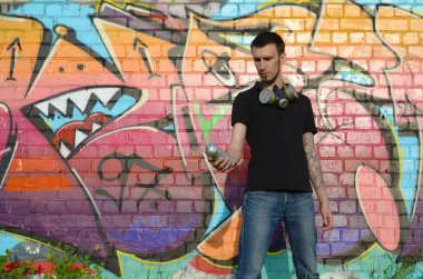 Beyaz beyaz grafiti sanatçısı siyah tişörtlü, sprey sprey boyalı tuğla duvarında renkli pembe grafitilerin yanında. Sokak sanatı ve çağdaş boyama süreci. Gençlik alt kültüründe eğlence