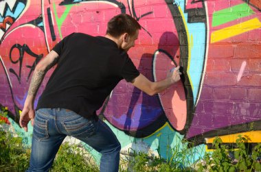 Sırt çantası ve gaz maskesi takan genç grafiti sanatçısı, tuğla duvara pembe tonlarda grafiti yapıyor. Sokak sanatı ve çağdaş boyama süreci. Gençlik alt kültüründe eğlence