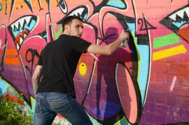 Sırt çantası ve gaz maskesi takan genç grafiti sanatçısı, tuğla duvara pembe tonlarda grafiti yapıyor. Sokak sanatı ve çağdaş boyama süreci. Gençlik alt kültüründe eğlence