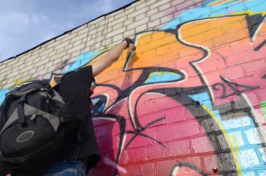 Sırt çantası ve gaz maskesi takan genç grafiti sanatçısı, tuğla duvara pembe tonlarda grafiti yapıyor. Sokak sanatı ve çağdaş boyama süreci. Gençlik alt kültüründe eğlence