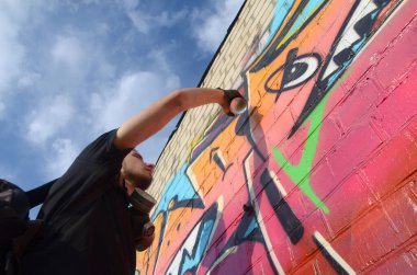 Sırt çantası ve gaz maskesi takan genç grafiti sanatçısı, tuğla duvara pembe tonlarda grafiti yapıyor. Sokak sanatı ve çağdaş boyama süreci. Gençlik alt kültüründe eğlence