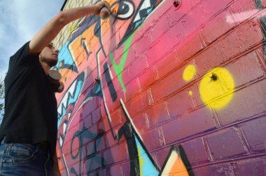 Sırt çantası ve gaz maskesi takan genç grafiti sanatçısı, tuğla duvara pembe tonlarda grafiti yapıyor. Sokak sanatı ve çağdaş boyama süreci. Gençlik alt kültüründe eğlence
