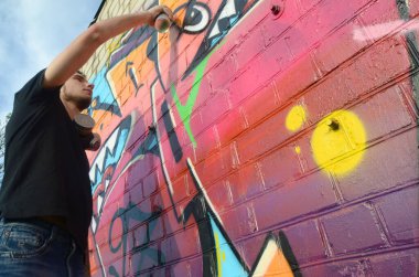 Sırt çantası ve gaz maskesi takan genç grafiti sanatçısı, tuğla duvara pembe tonlarda grafiti yapıyor. Sokak sanatı ve çağdaş boyama süreci. Gençlik alt kültüründe eğlence