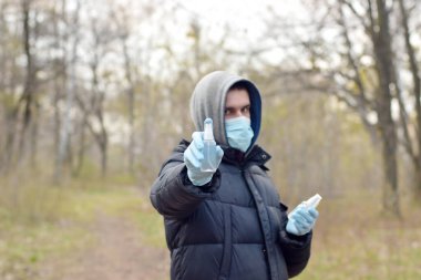 Koruyucu maskeli genç adam dezenfektan sprey şişelerini dışarıda, yay ağacında gösteriyor. Karantina sırasında sıvı antiseptik sprey kullanımı kavramı