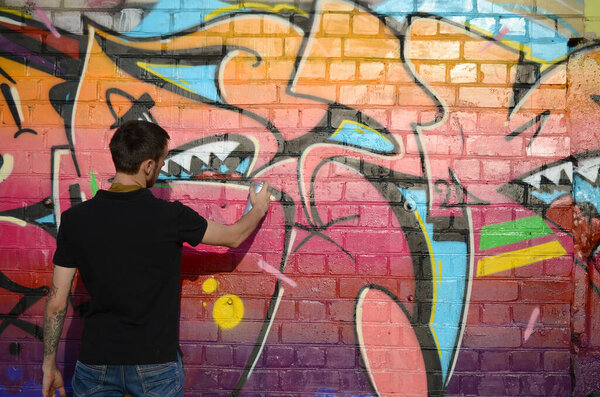 Young graffiti artist with backpack and gas mask on his neck paints colorful graffiti in pink tones on brick wall. Street art and contemporary painting process. Entertainment in youth subculture