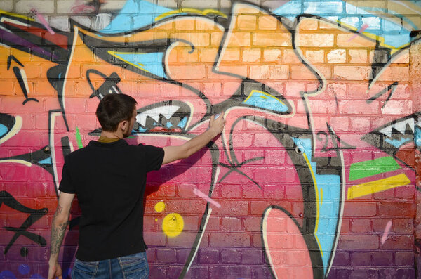 Young graffiti artist with backpack and gas mask on his neck paints colorful graffiti in pink tones on brick wall. Street art and contemporary painting process. Entertainment in youth subculture