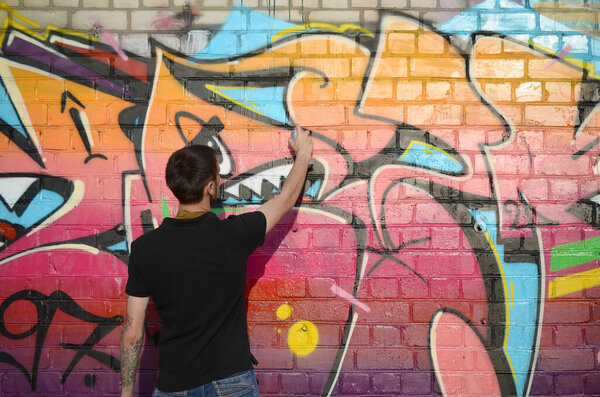 Young graffiti artist with backpack and gas mask on his neck paints colorful graffiti in pink tones on brick wall. Street art and contemporary painting process. Entertainment in youth subculture