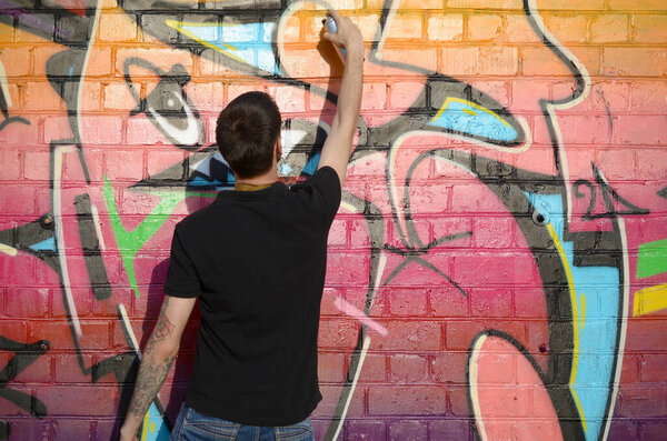 Young graffiti artist with backpack and gas mask on his neck paints colorful graffiti in pink tones on brick wall. Street art and contemporary painting process. Entertainment in youth subculture