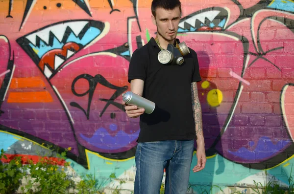 Young caucasian graffiti artist in black t-shirt with aerosol spray can near colorful pink graffiti on brick wall. Street art and contemporary painting process. Entertainment in youth subculture