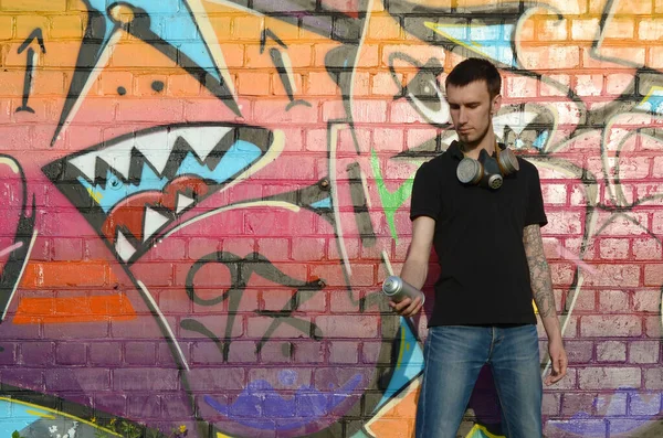 Young caucasian graffiti artist in black t-shirt with aerosol spray can near colorful pink graffiti on brick wall. Street art and contemporary painting process. Entertainment in youth subculture