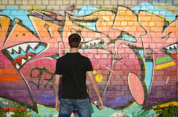 Young graffiti artist with backpack and gas mask on his neck paints colorful graffiti in pink tones on brick wall. Street art and contemporary painting process. Entertainment in youth subculture