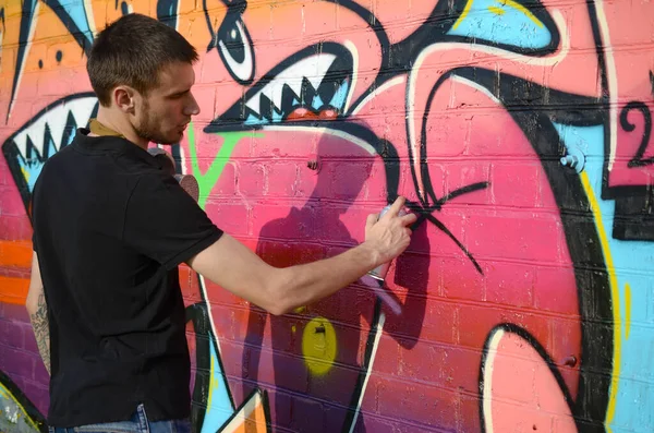 Young graffiti artist with backpack and gas mask on his neck paints colorful graffiti in pink tones on brick wall. Street art and contemporary painting process. Entertainment in youth subculture