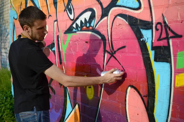 Young graffiti artist with backpack and gas mask on his neck paints colorful graffiti in pink tones on brick wall. Street art and contemporary painting process. Entertainment in youth subculture