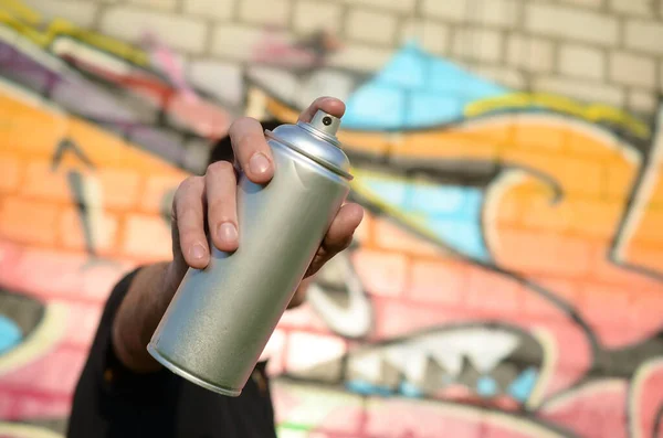 Young graffiti artist aims his spray can on background of colorful graffiti in pink tones on brick wall. Street art and contemporary painting process. Entertainment in youth subculture