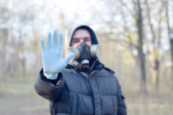 Blurred portrait of young man in protective gas mask and rubber disposable gloves shows stop gesture outdoors in spring wood. Concept of protective goods usage during quarantine