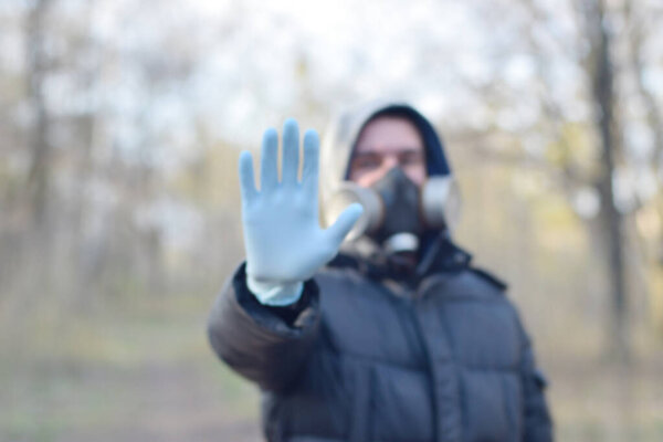 Blurred portrait of young man in protective gas mask and rubber disposable gloves shows stop gesture outdoors in spring wood. Concept of protective goods usage during quarantine