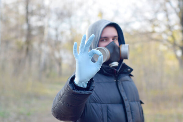 Portrait of young man in protective gas mask and rubber disposable gloves shows okay gesture outdoors in spring wood. Concept of protective goods usage during quarantine