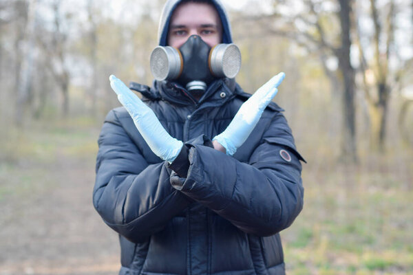 Portrait of young man in protective gas mask and rubber disposable gloves shows stop gesture outdoors in spring wood. Concept of protective goods usage during quarantine
