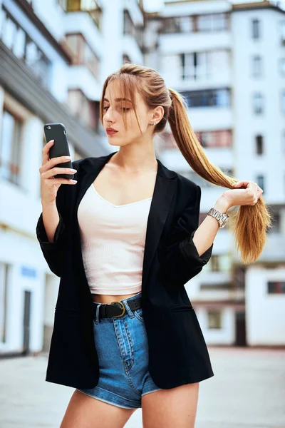 Elegante ragazza bionda in gonna di tulle e camicia di jeans con labbra viola si sta divertendo e fa selfie con un viso bacio e con il segno della vittoria dita con strisce di legno marrone balks dietro . — Foto Stock