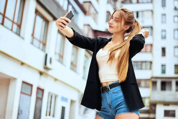 Snygg blond tjej i tyll kjol och denim skjorta med lila läppar är att ha kul och gör selfie med en kyss ansikte och med seger tecken fingrar med randig brun trä hinder bakom. — Stockfoto