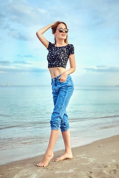 Mujer elegante con el pelo largo de pie en blusa y pantalones cortos vaqueros. Hermosa chica caminando en la playa al atardecer —  Fotos de Stock