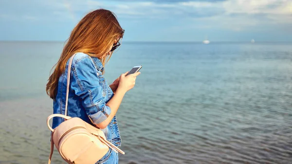 Una ragazza turistica cammina lungo il mare con uno zaino, in cianfrusaglie e occhiali da sole. Con un telefono cellulare, scatta foto su uno smartphone. Selfie — Foto Stock