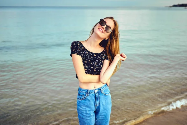 Donna elegante con i capelli lunghi in piedi in camicetta e pantaloncini jeans. Bella ragazza che cammina in spiaggia al tramonto — Foto Stock