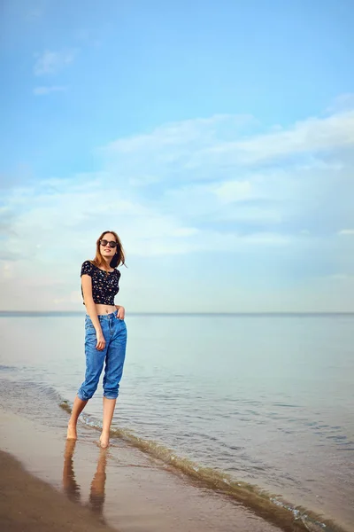 Elegantní žena s dlouhými vlasy stojící v halence a jeans kraťasy. Krásná dívka procházky na pláži při západu slunce — Stock fotografie