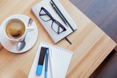 Bir fincan kahve bir fincan tabağı üzerinde bir not defteri ve kalem ve gözlük yanındaki ahşap bir masa üzerinde bir kaşıkla