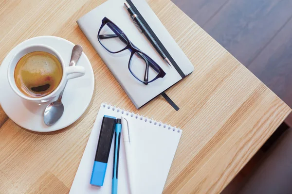 Bir fincan kahve bir fincan tabağı üzerinde bir not defteri ve kalem ve gözlük yanındaki ahşap bir masa üzerinde bir kaşıkla