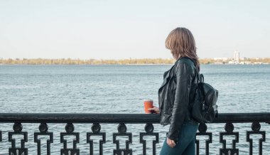  Kız bir deri ceket ve kot bir sırt çantası ile bir bardak kahve ile sahildeki çitin yanında duruyor ve nehirde görünüyor