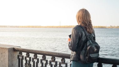  Kız bir deri ceket ve kot bir sırt çantası ile bir bardak kahve ile sahildeki çitin yanında duruyor ve nehirde görünüyor