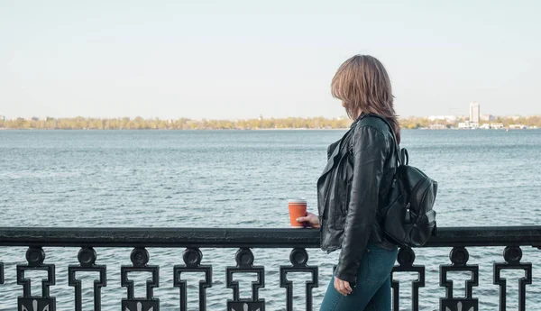  Kız bir deri ceket ve kot bir sırt çantası ile bir bardak kahve ile sahildeki çitin yanında duruyor ve nehirde görünüyor