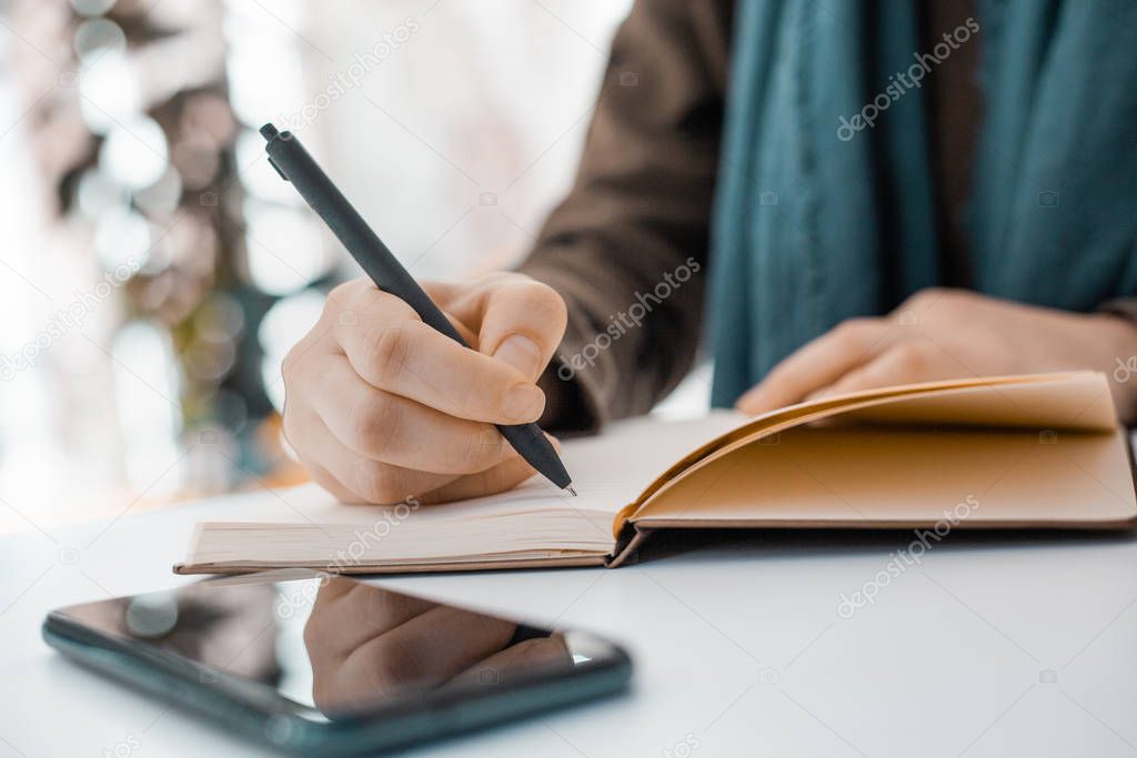 girl writes in a notebook with a black pen next to the phone on a white table
