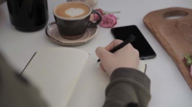 Bir kızın eli seni sevdiğimi beyaz bir masanın üzerinde bir fincan kahve ve bir telefonun yanında yazıyor.