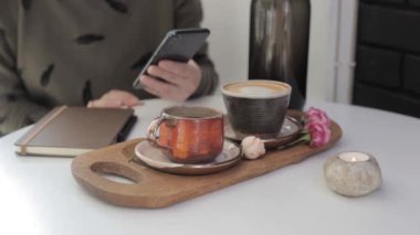 Elinde telefon tutan ve sayfaları kaydıran bir kız kahve ve günlükle masada oturuyor.