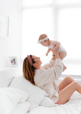 Mutlu bir aile sabahı, evde pijamalı genç bir anne evdeki rahat bir yatakta oynuyor, çocuğunu öpüyor ve sarılıyor.