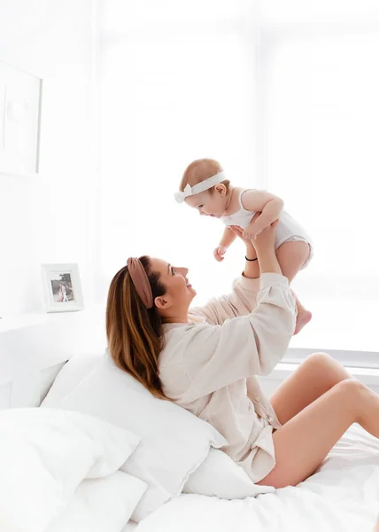 Mutlu bir aile sabahı, evde pijamalı genç bir anne evdeki rahat bir yatakta oynuyor, çocuğunu öpüyor ve sarılıyor.