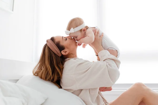 Mutlu bir aile sabahı, evde pijamalı genç bir anne evdeki rahat bir yatakta oynuyor, çocuğunu öpüyor ve sarılıyor.