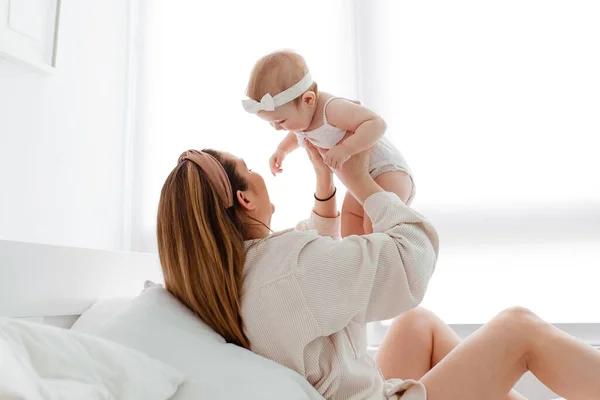 Mutlu bir aile sabahı, evde pijamalı genç bir anne evdeki rahat bir yatakta oynuyor, çocuğunu öpüyor ve sarılıyor.